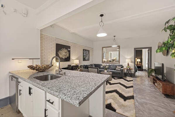 A modern kitchen with granite countertops, a built-in dishwasher, and a dining area with a gray sofa and a coffee table.