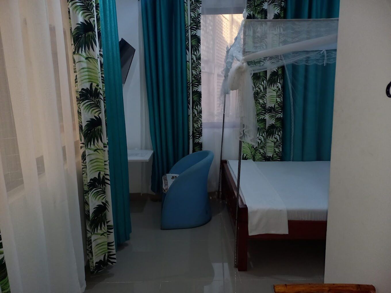 A small, narrow room with a bed, a chair, and curtains with a tropical leaf pattern.