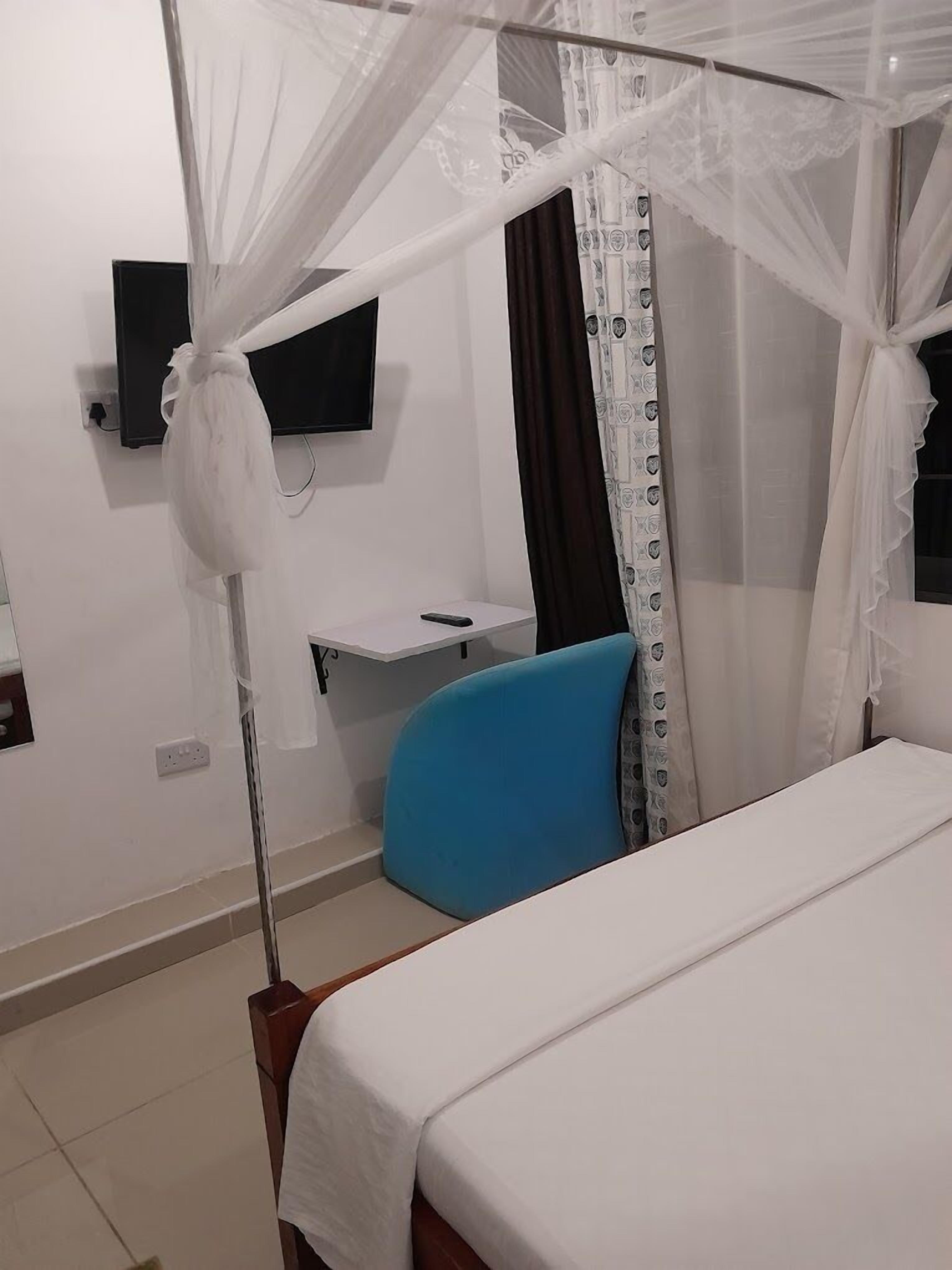 Una camera d'albergo con un letto, una poltrona blu, una TV a parete e un comodino.