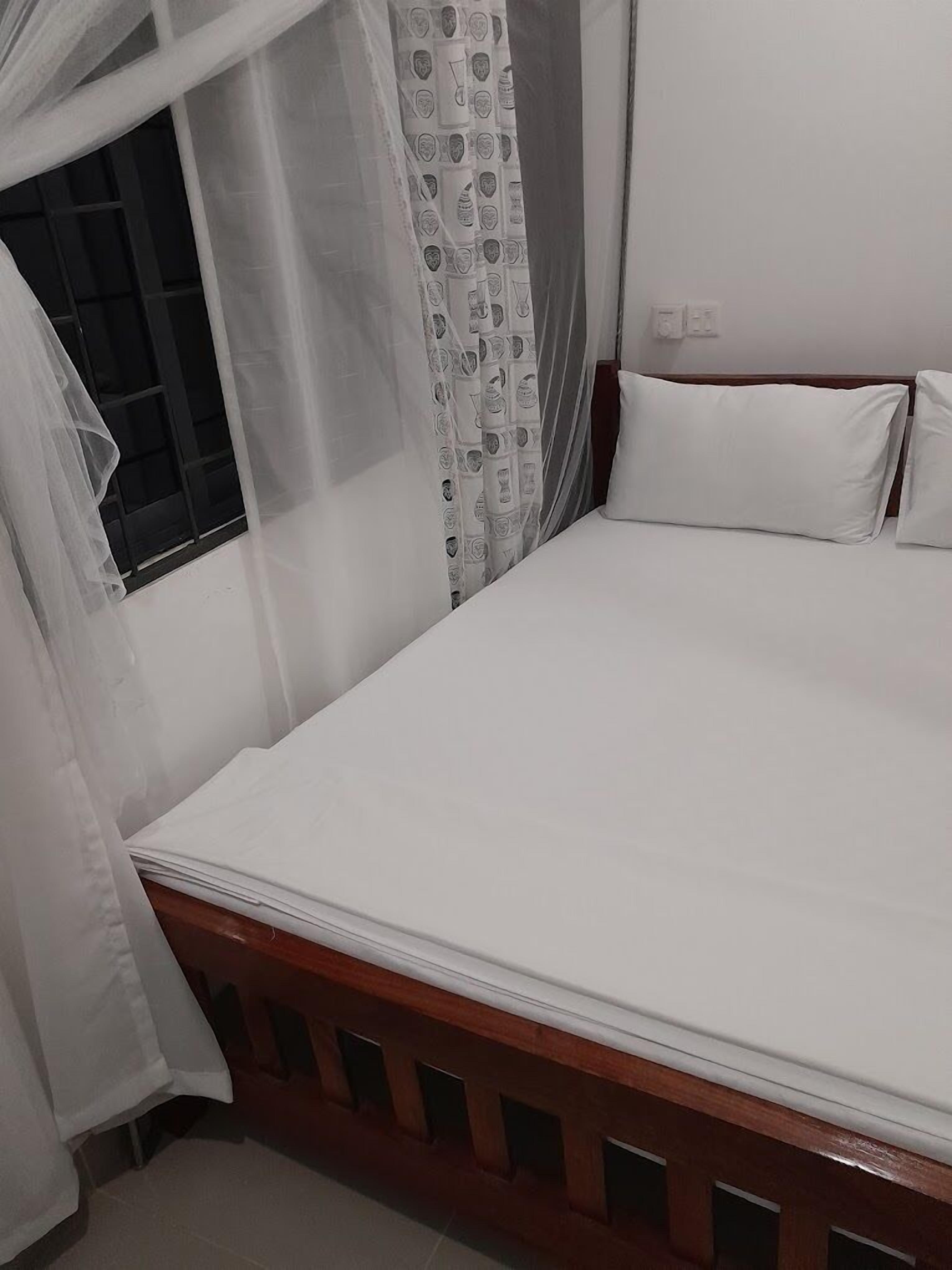Un dormitorio con estructura de cama de madera, ropa de cama blanca, una ventana con cortina y un enchufe de pared.