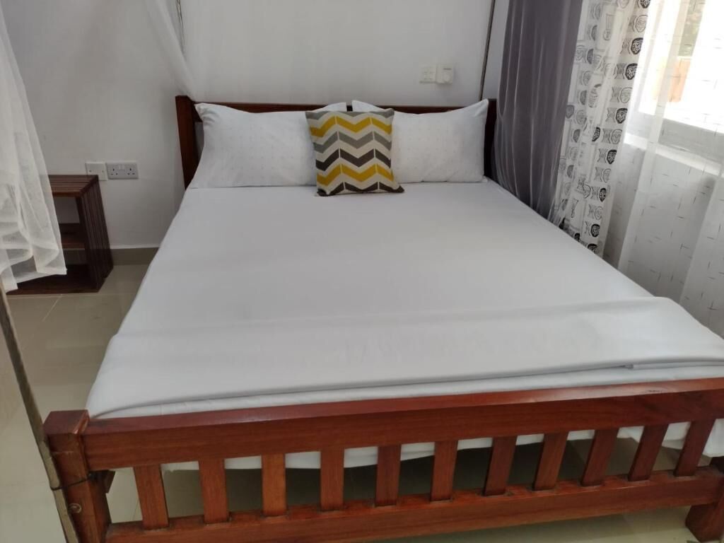 Una cama con estructura de madera, ropa de cama blanca y almohada estampada.