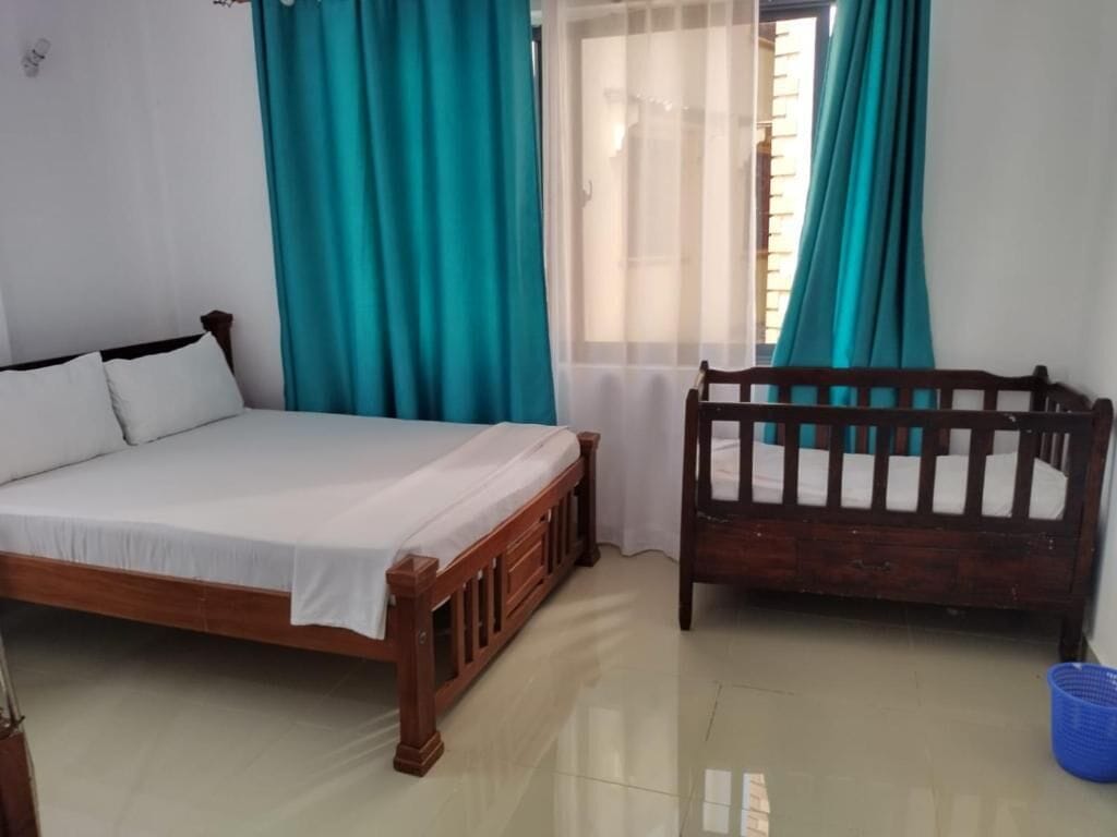 A bedroom with a wooden bed, a wooden crib, teal curtains, and a window with blinds.