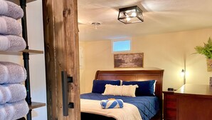 A bedroom with a wooden bed, a nightstand, a wall-mounted shelf with towels, a ceiling light, and a window with blinds. - Hot Tub Outdoor Living Game Room Coffee Bar (South Bend)