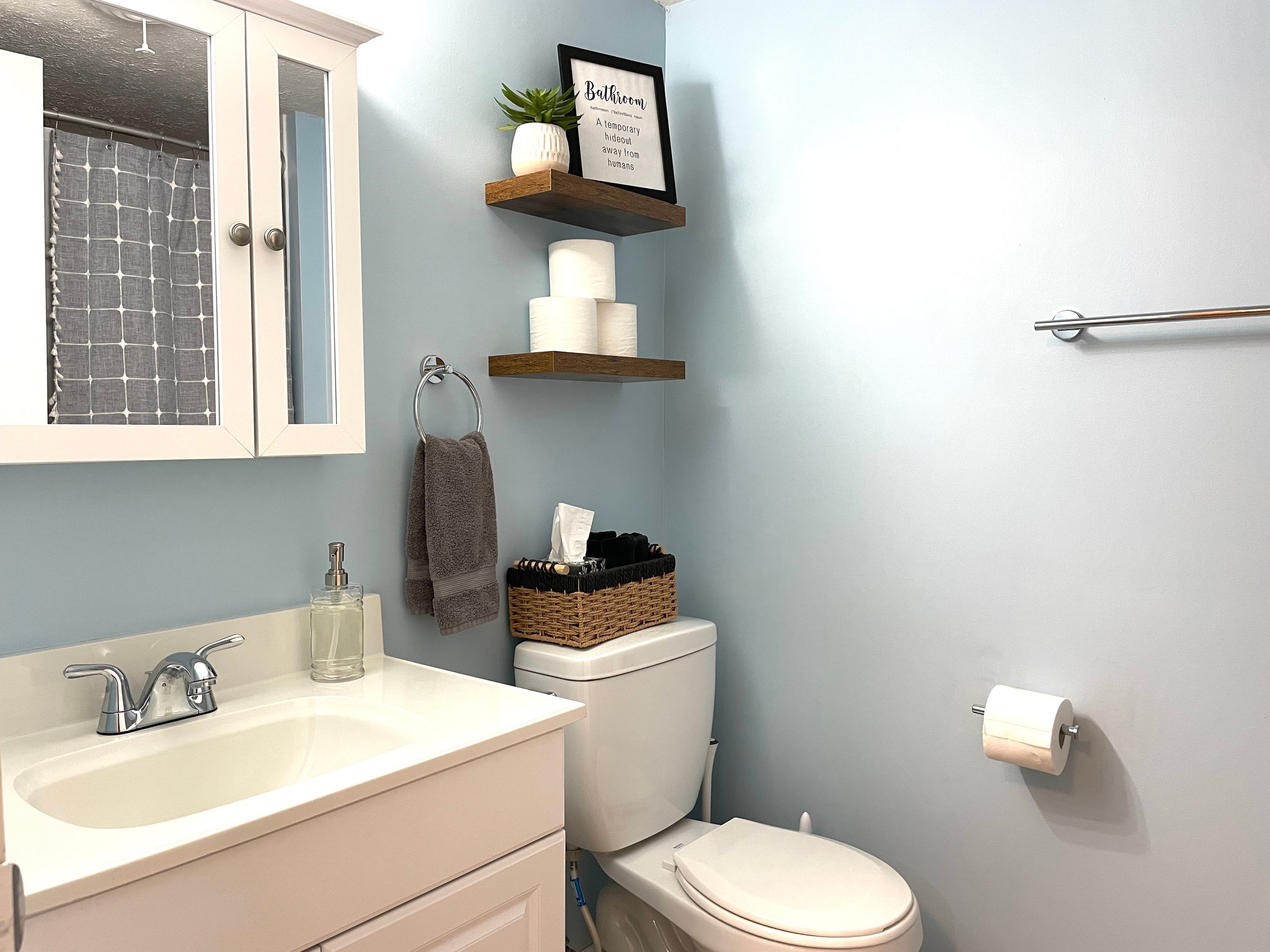 Baño con lavamanos blanco, inodoro y cortina de ducha. Se observan repisas con artículos de aseo y una cita enmarcada en la pared.