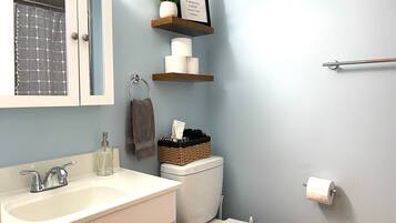 Baño con lavamanos blanco, inodoro y cortina de ducha. Se observan repisas con artículos de aseo y una cita enmarcada en la pared.