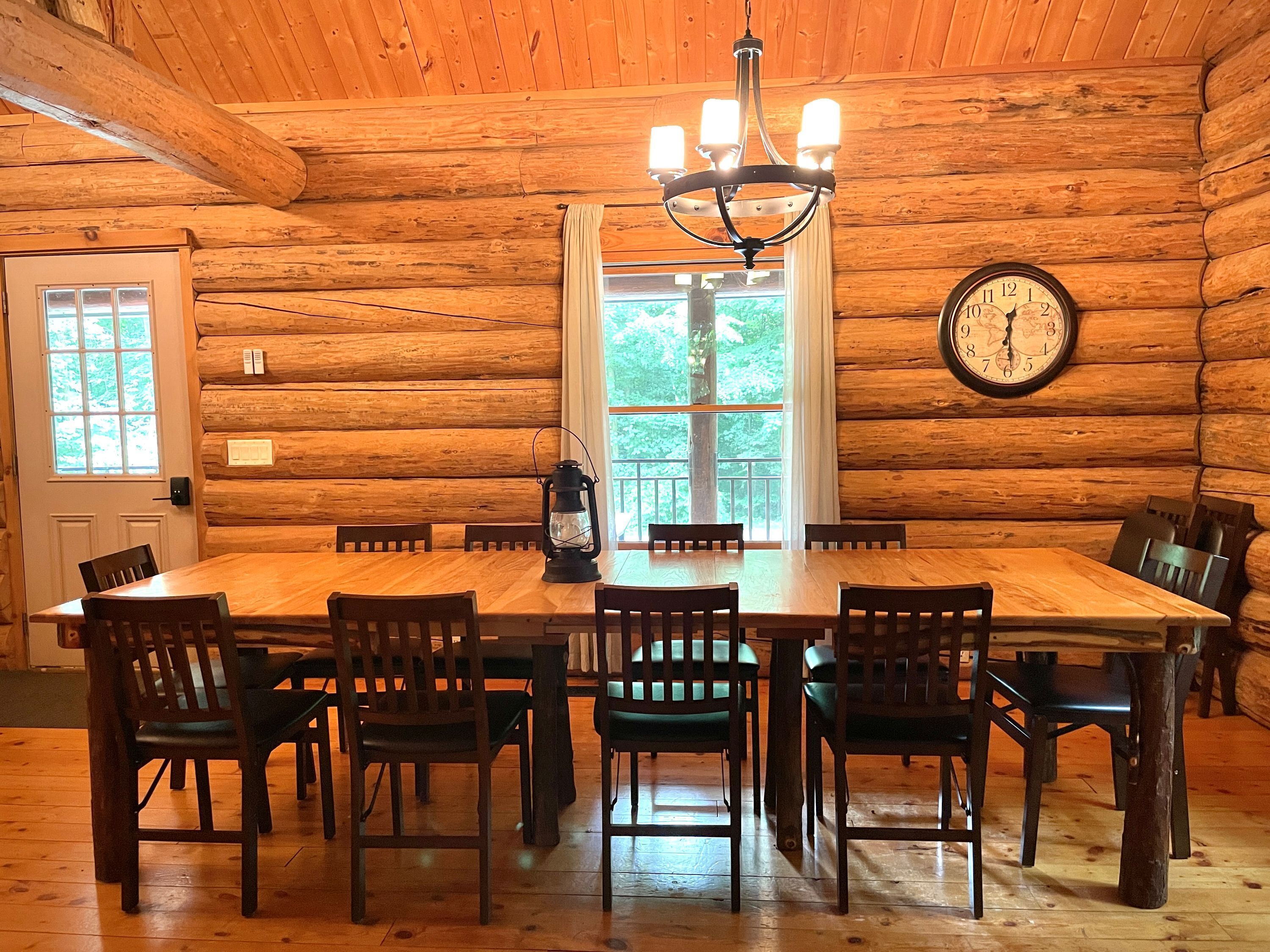 A log cabin dining room with a wooden table and chairs, a chandelier, a clock, and a window with curtains.