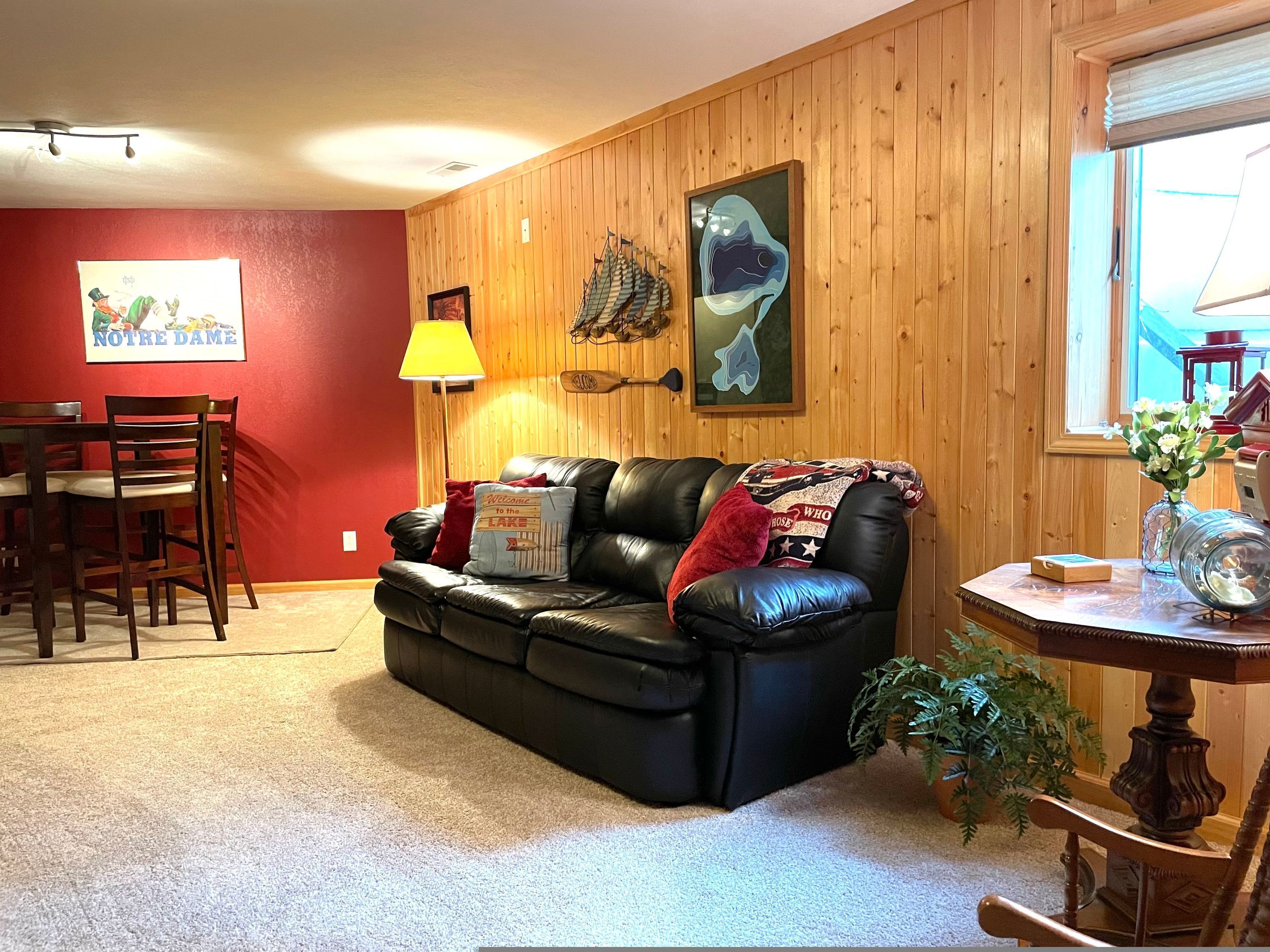 A living room with a black leather couch, wooden walls, a dining table with chairs, a lamp, and a window with a view.