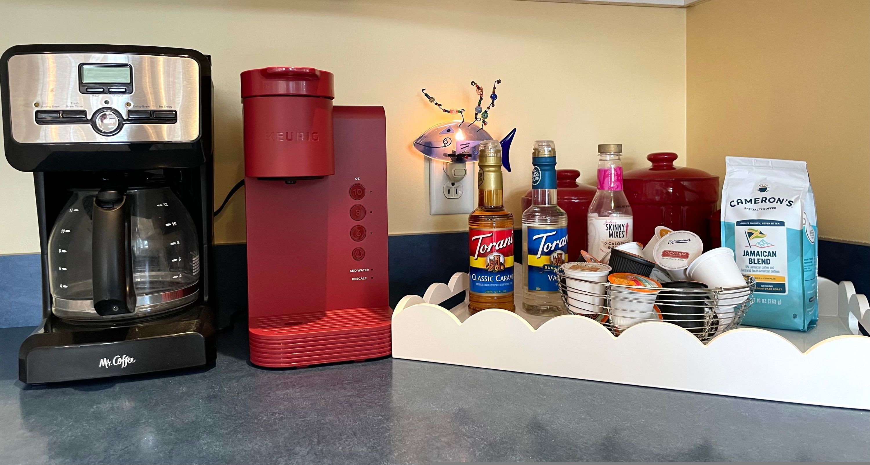 Kaffestation med en Keurig-kaffemaskine, en rød kaffedispenser samt diverse kaffeprodukter og -beholdere.