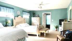 Une chambre à coucher comprenant un lit, une table de chevet, une commode, une chaise, un ventilateur de plafond et une porte donnant sur une autre pièce.