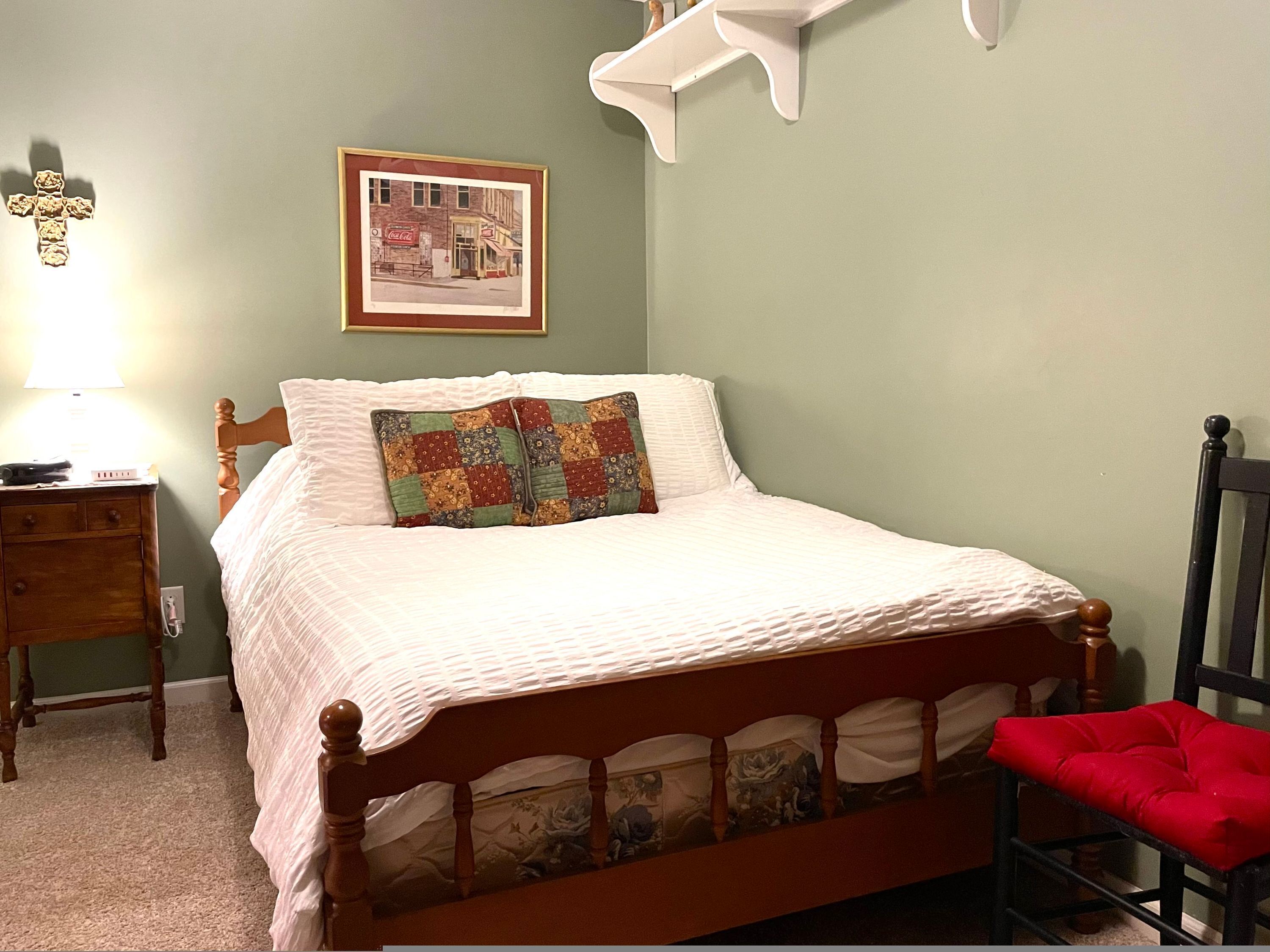 Una cama bien hecha con una colcha blanca acolchada y un cojín colorido de patchwork. Una mesita de noche de madera con una lámpara y una pequeña mesa está al lado de la cama. Un cuadro enmarcado cuelga de la pared sobre la cama.
