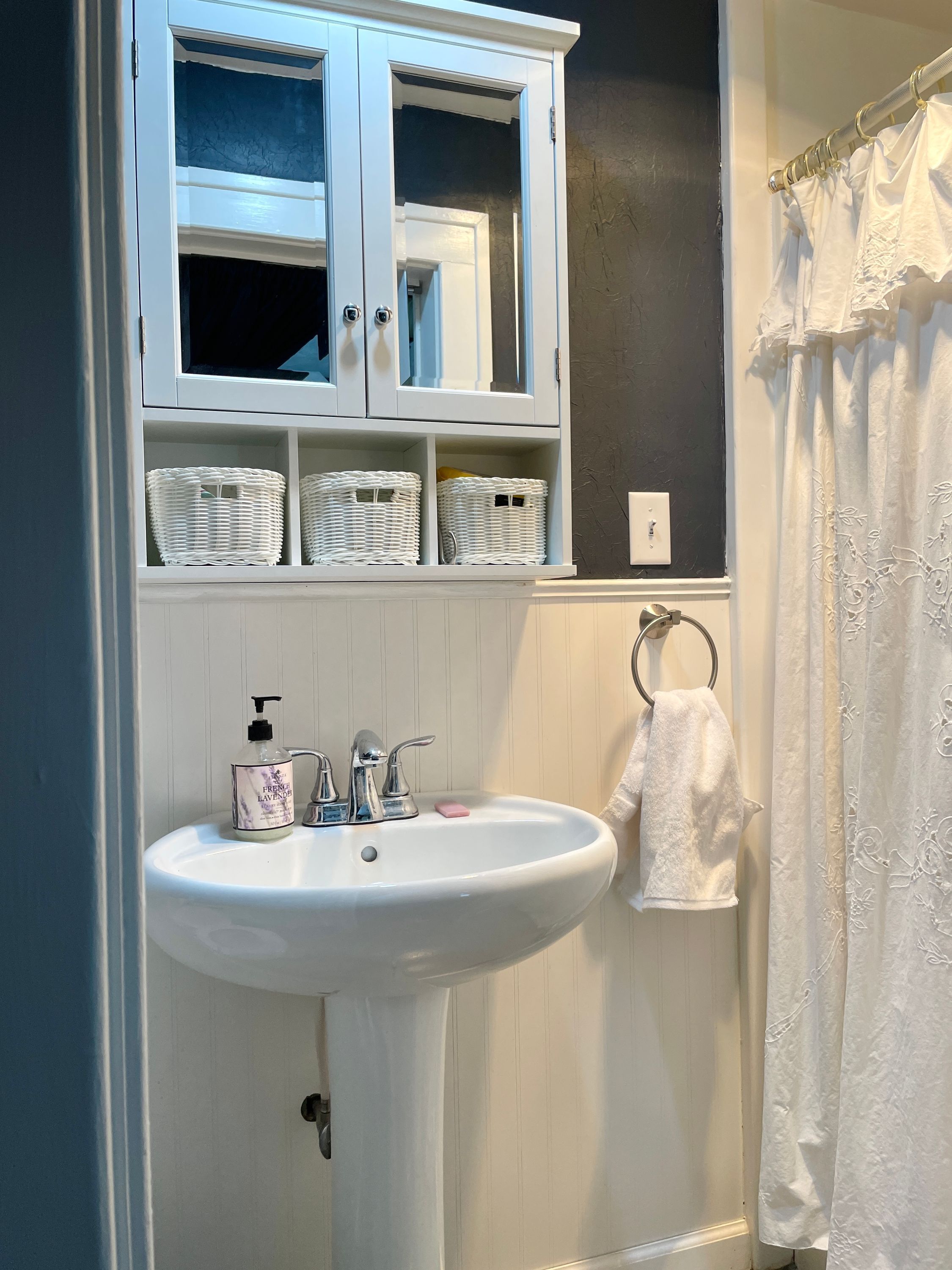 Un baño con un lavamanos blanco, un mueble con cestas de mimbre, un perchero y una cortina de ducha.