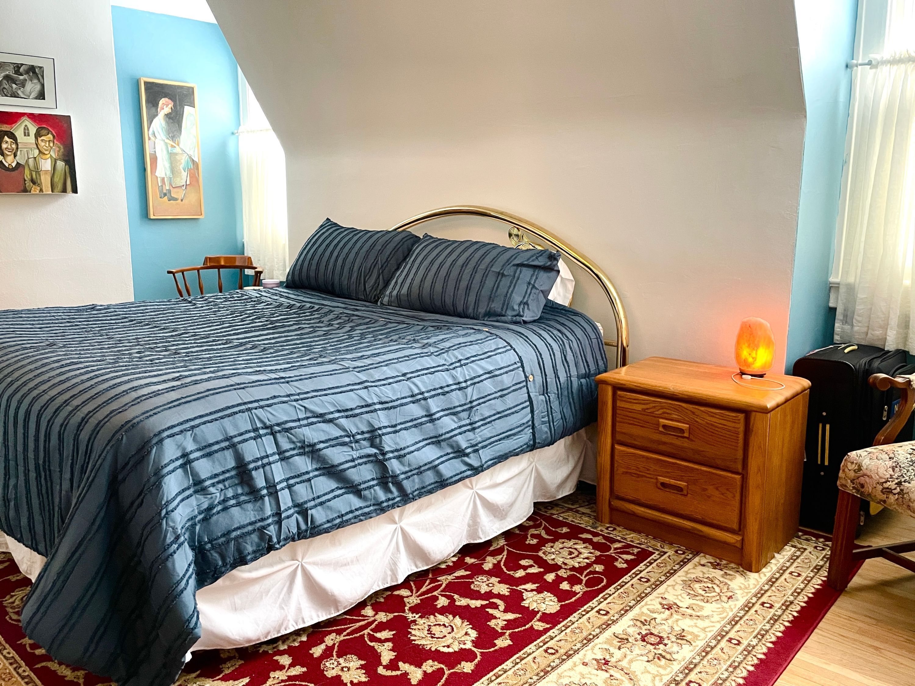 A bedroom with a bed, bedside table, lamp, chair, and a red and white patterned rug.