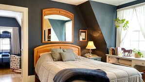 Une chambre à coucher avec un grand lit, une tête de lit en bois, une table de chevet avec une lampe, un miroir, une fenêtre avec des rideaux, une bibliothèque et une plante en pot.