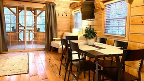 A wooden cabin interior with a dining table, chairs, a potted plant, and a large window with blinds. - Secluded Shipshewana Log Cabin In the Woods (Shipshewana)