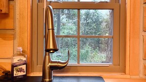 A kitchen with a stainless steel faucet, a bottle of lemon drop, and a window with a view of trees. - Secluded Shipshewana Log Cabin In the Woods (Shipshewana)