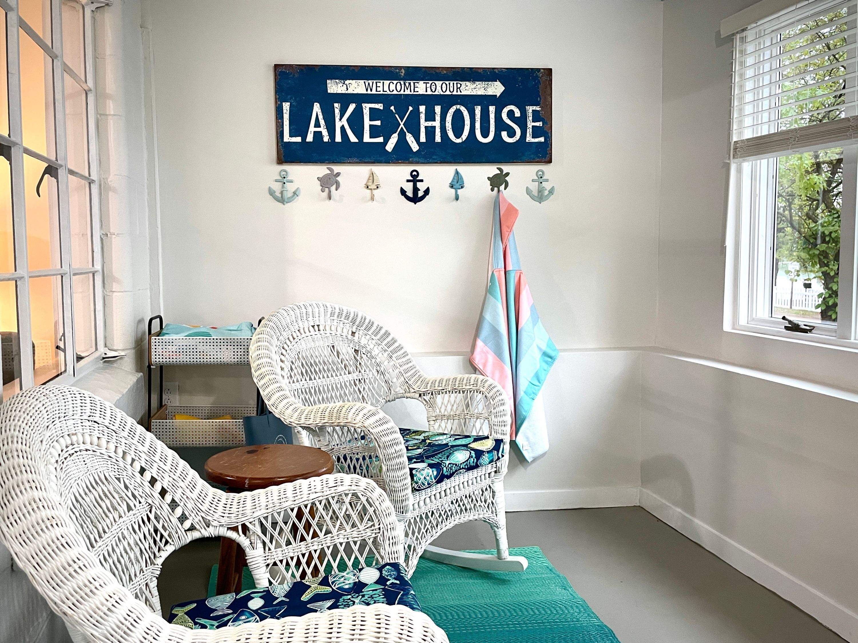 Quarto com cadeiras de vime, uma pequena mesa, uma placa com a inscrição 'Bem-vindos à nossa Casa de Lago' e decorações náuticas.