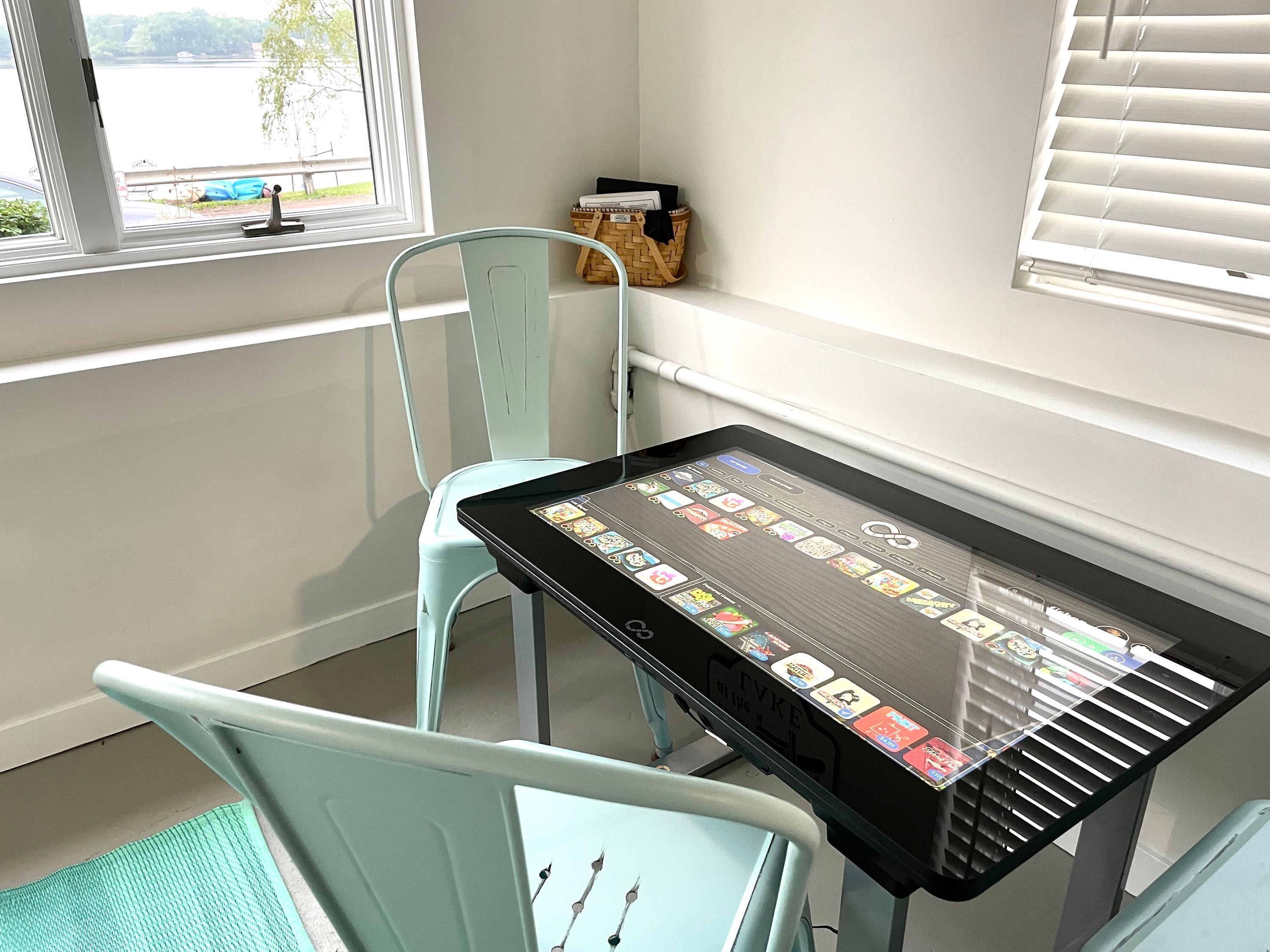 A room with a table displaying a digital interface, a window with a view of a body of water, and a wicker basket on a shelf.