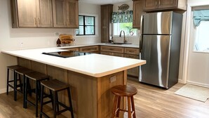A modern kitchen with wooden cabinets, a stainless steel refrigerator, a large island with stools, and a window with blinds. - Lake Front Cottage Kayaks Game Table Fun (Shipshewana)
