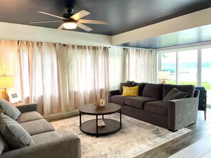 Ein Wohnzimmer mit Deckenventilator, einer Couch, einem Couchtisch und Blick ins Freie durch große Fenster.