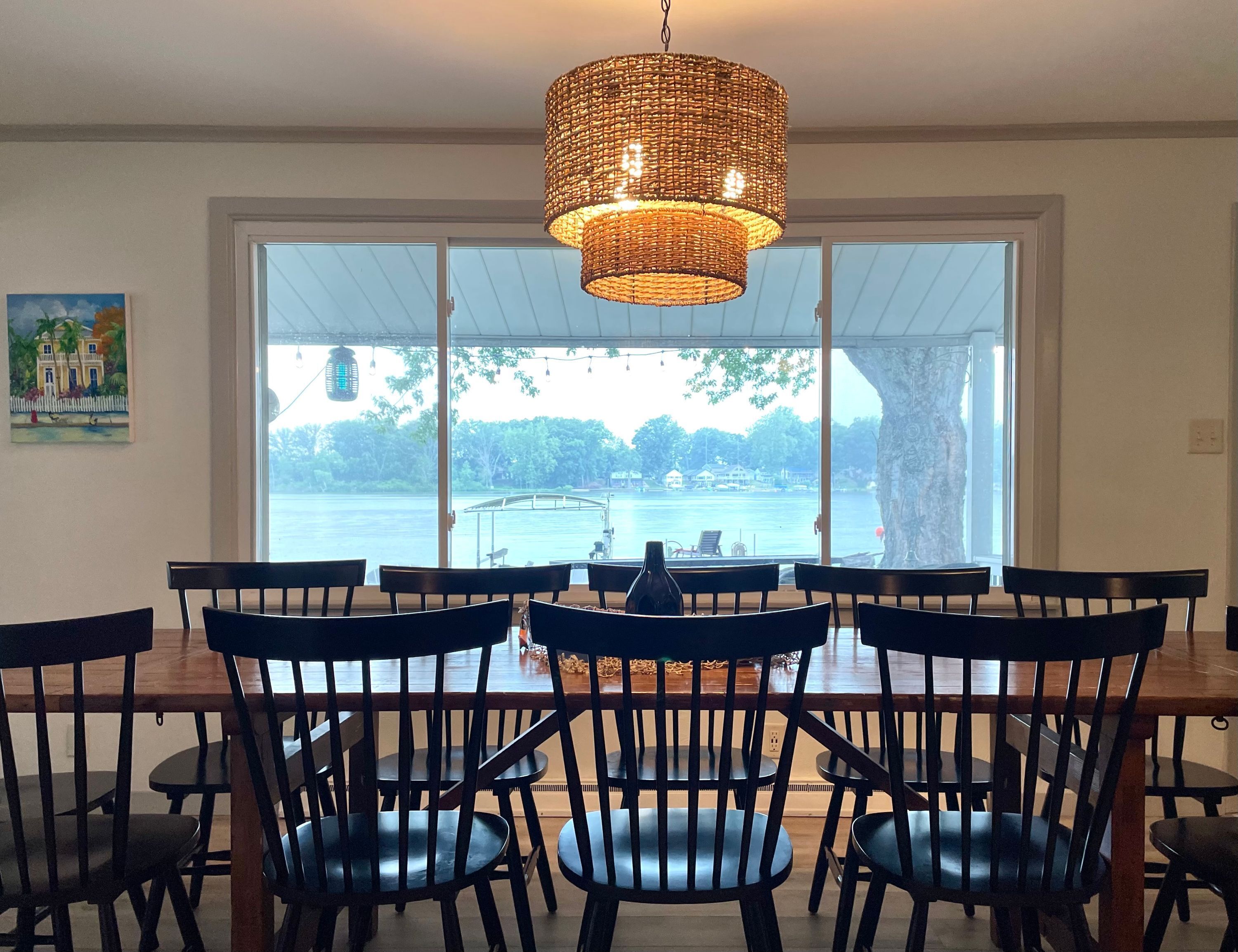 Un espace repas comprenant une grande table et des chaises, un luminaire en fibres tressées et une vue sur un plan d’eau grâce à de grandes fenêtres.