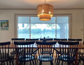 A dining area with a large table and chairs, a woven light fixture, and a view of a body of water through large windows. - Lake House Kayaks Patio Deck Private Pier (Rome City)