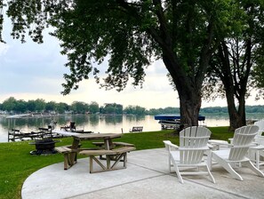 Eine Uferterrasse mit Picknicktischen und Adirondack-Stühlen mit Blick auf einen ruhigen See, im Hintergrund Boote und ein Steg.