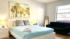 A bedroom with a bed, bedside tables, a nightstand, a dark sofa, a window with blinds, and a painting of palm trees. - Sarasota Family Home Private Heated Pool Relax (Sarasota)