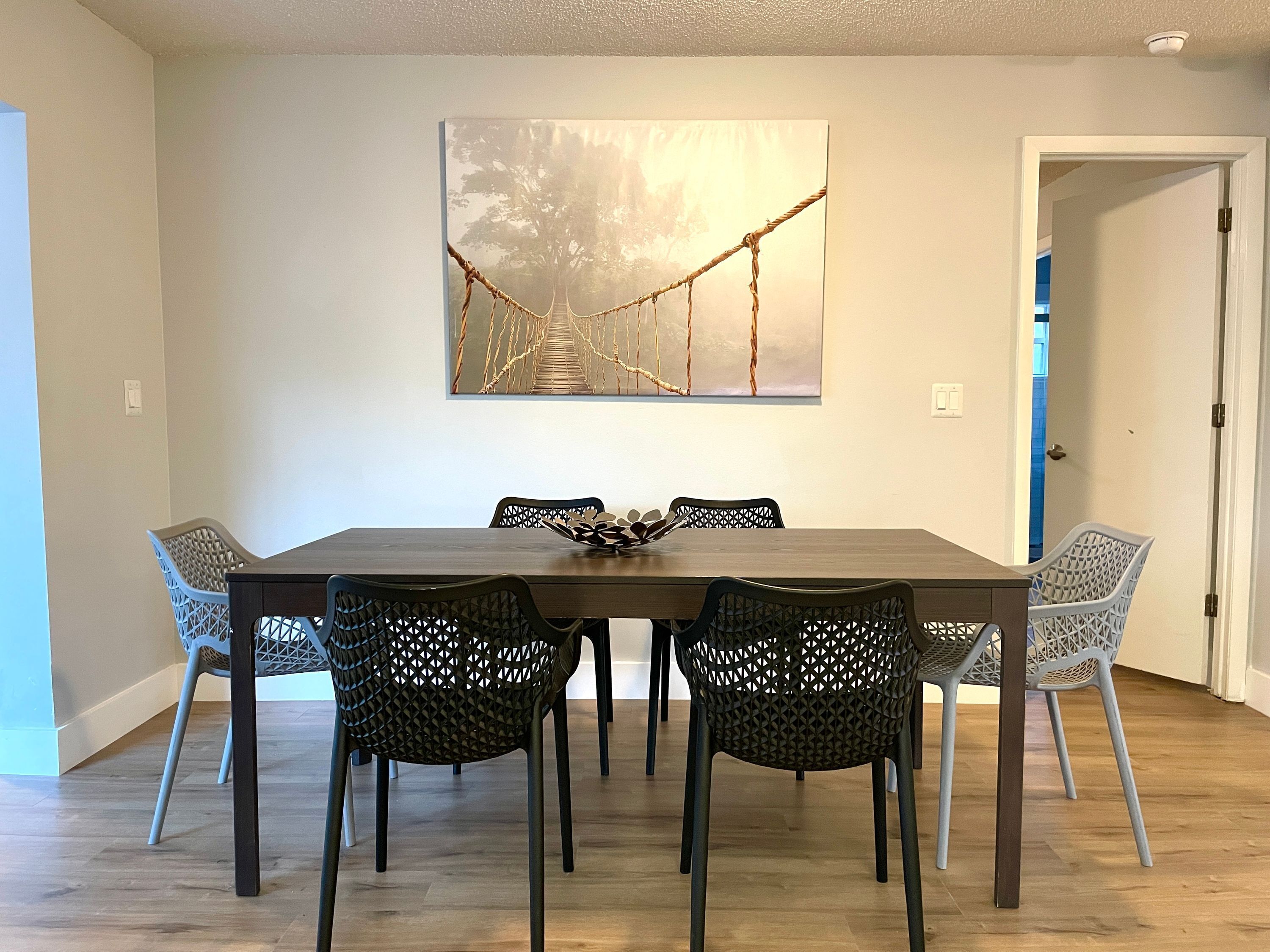 A dining area with a wooden table and six black chairs with a geometric pattern. A large abstract painting hangs on the wall above the table.