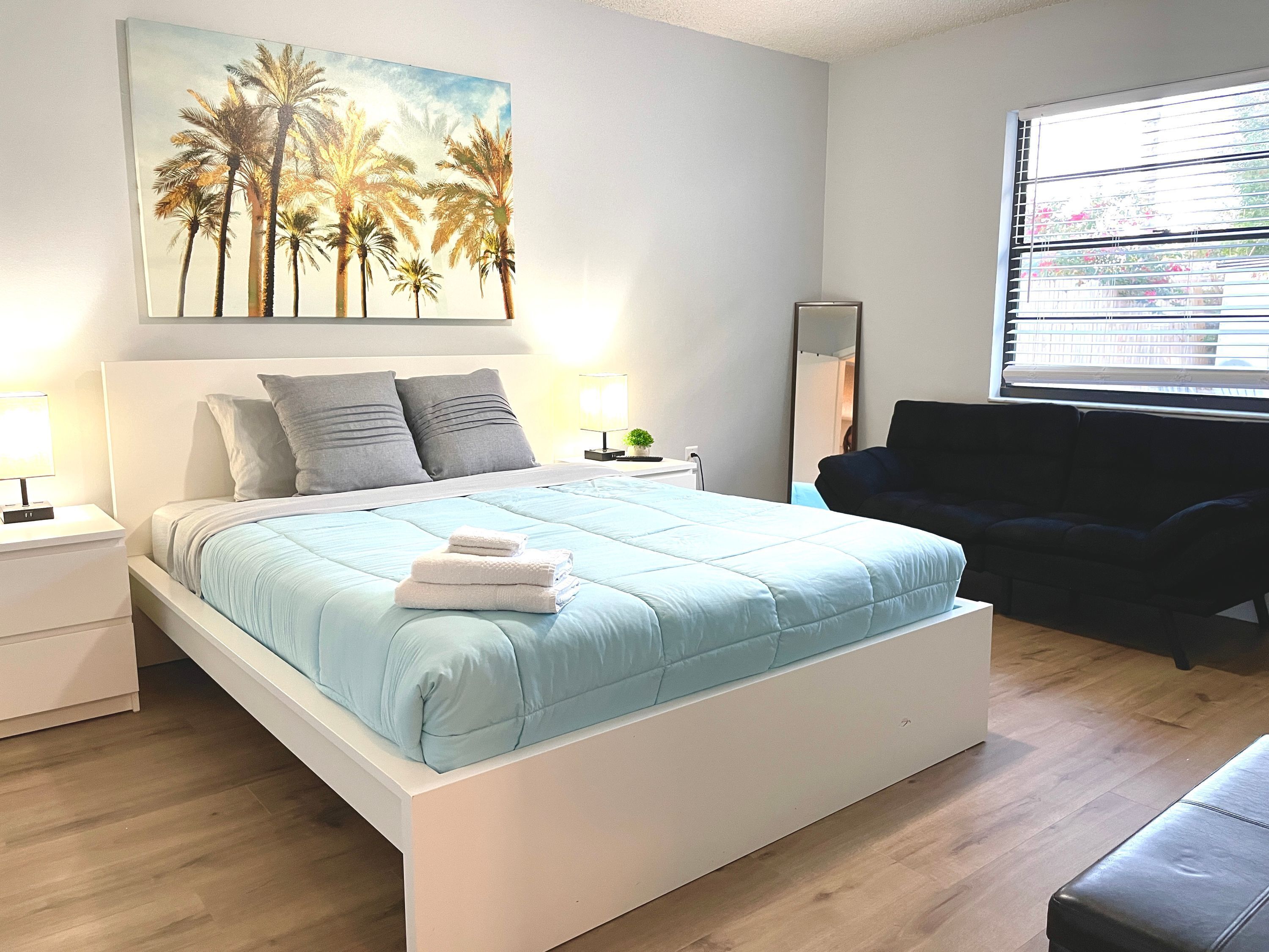 Un dormitorio con cama, mesitas de noche, una silla, una ventana y un cuadro con palmeras.