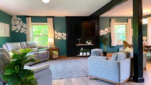 Un salon moderne avec un canapé, un fauteuil et un téléviseur fixé à un mur noir. On y trouve également des plantes en pot et un tapis à motifs sur le sol.