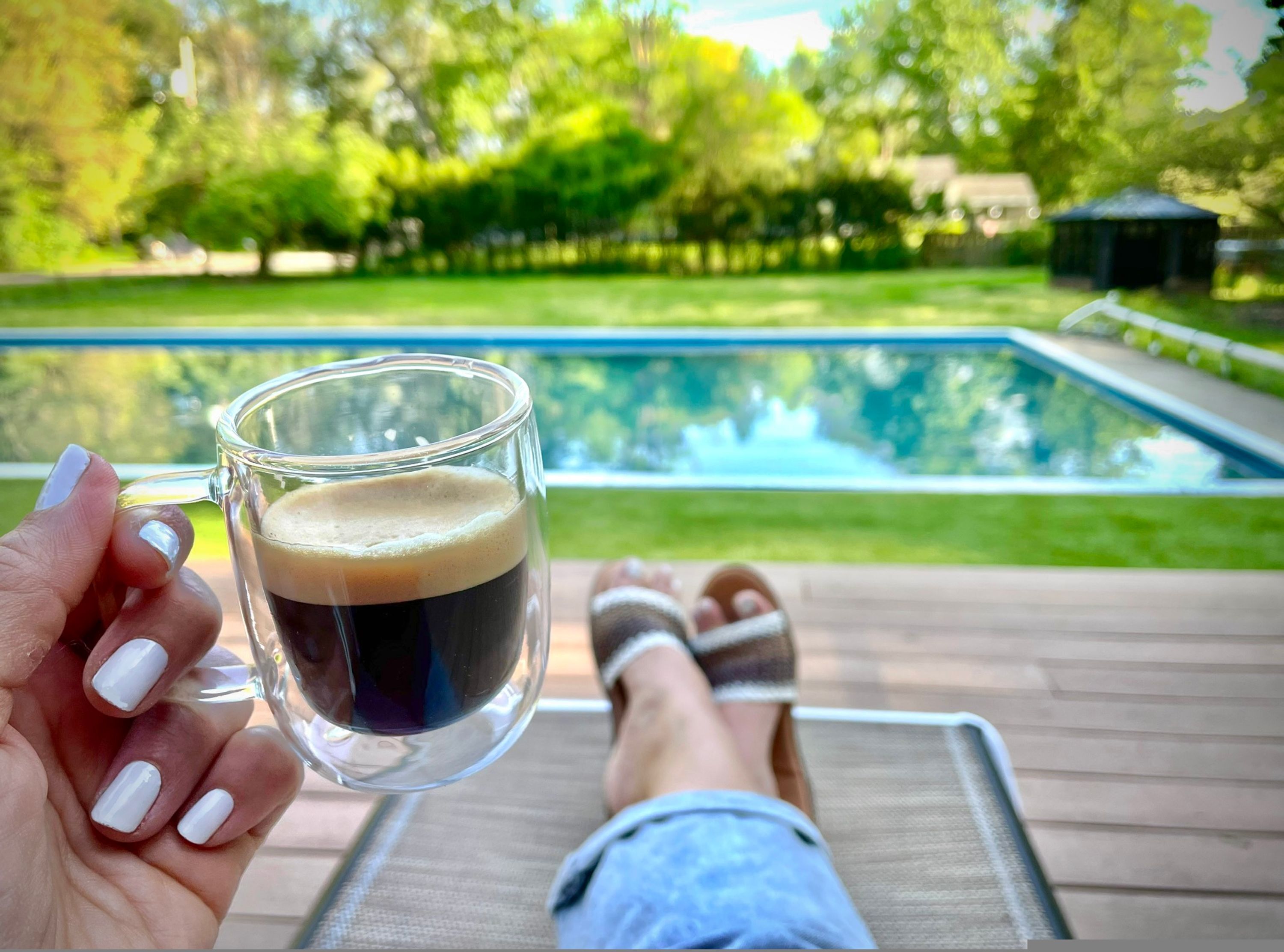 Eine Person hält ein Glas mit dunkler Flüssigkeit, vermutlich Kaffee, auf einer Terrasse mit Blick auf einen Swimmingpool.
