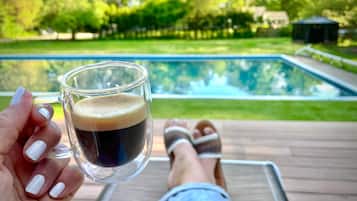 Une personne tenant un verre contenant un liquide sombre, vraisemblablement du café, sur une terrasse donnant sur une piscine.