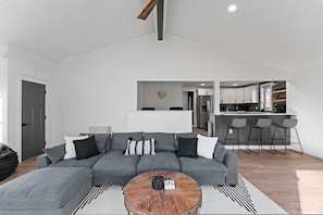 A modern living room with a grey sectional sofa, a wooden coffee table, and a kitchen area in the background. - Pool Hot Tub 5 min to ND Coffee Bar Games (South Bend)
