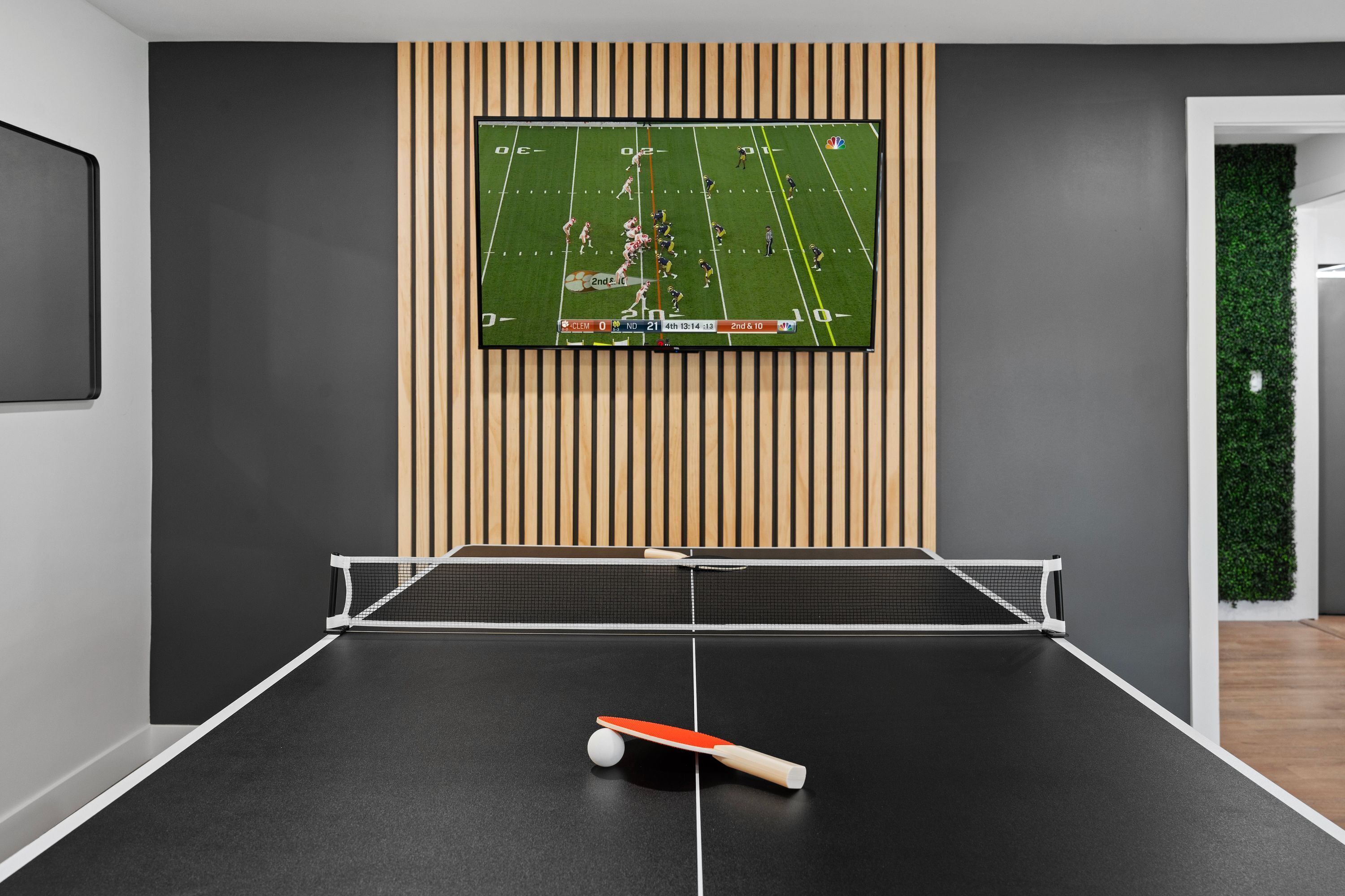 Ett bordtennisbord med en tv-skärm som visar en fotbollsmatch, en vägg med träpaneler och en grön vägg med en växt.