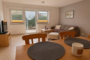 A living room with a sofa, a television, a dining table with placemats, and a window with curtains. - Apartment with terrace in Hollersbach (Hollersbach im Pinzgau)