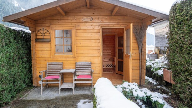 Una cabaña de madera con dos sillas y un banco en el exterior, una puerta que da al interior, y nieve en el suelo.