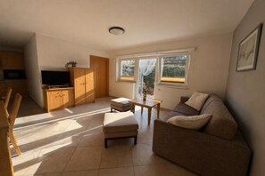 A living room with a sofa, ottoman, TV, wooden cabinets, and a window with a view. - Apartment with terrace in Hollersbach (Hollersbach im Pinzgau)