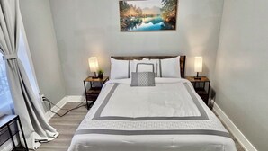 Una cama bien hecha con un edredón blanco y gris, dos lámparas de noche y un cuadro de un lago en la pared.