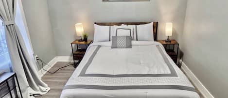 A neatly made bed with a white and gray bedspread, two bedside lamps, and a painting of a lake on the wall.