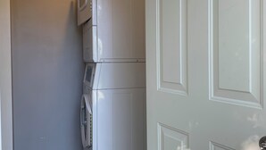 Lavandería con lavadora y secadora apiladas, piso de baldosas y una puerta con manija.