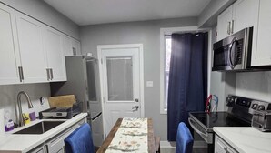 Una cocina compacta con armarios blancos, un refrigerador, un microondas y una estufa. Hay una mesa de comedor con sillas azules y un mantel.