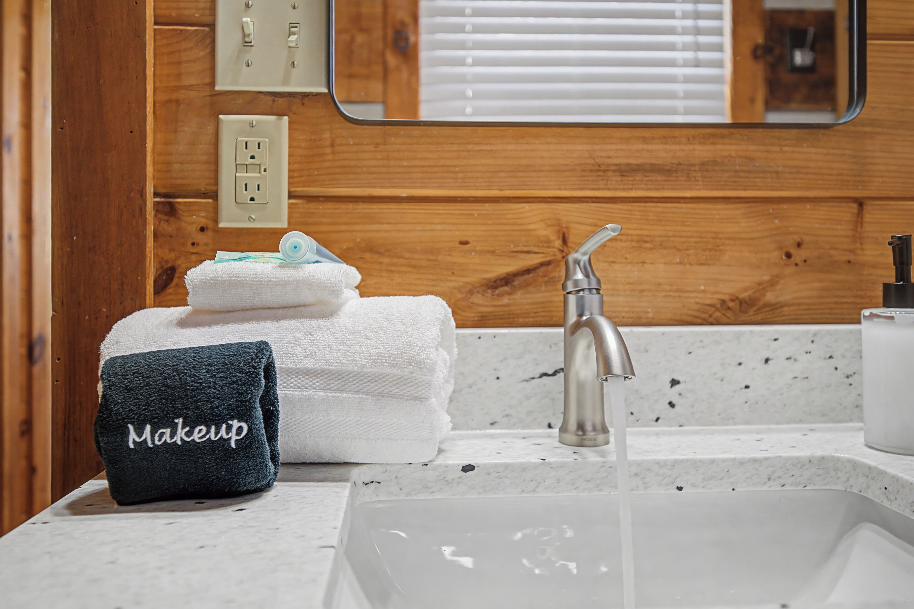Une salle de bain avec un mur en bois, un lavabo blanc doté d ’un robinet moderne, et un porte-serviettes sur lequel sont posés une serviette blanche et une serviette bleu foncé ornée d’une broderie représentant des produits de maquillage.