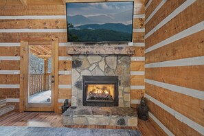 L’intérieur d’une cabane en rondins, avec une cheminée en pierre, un téléviseur fixé au-dessus et une porte en bois donnant sur l’extérieur.