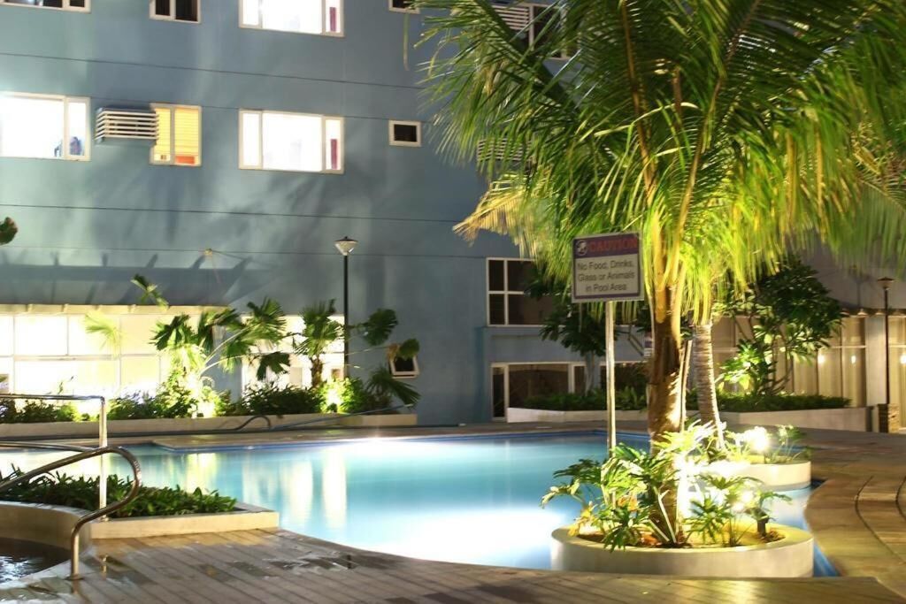 A swimming pool area with a blue building in the background, surrounded by palm trees and plants.