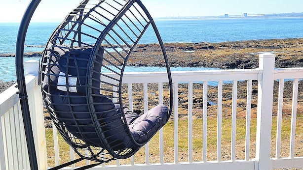 Een hangstoel op een houten terras met een witte leuning, met uitzicht op een rotsachtige kustlijn en de oceaan.
