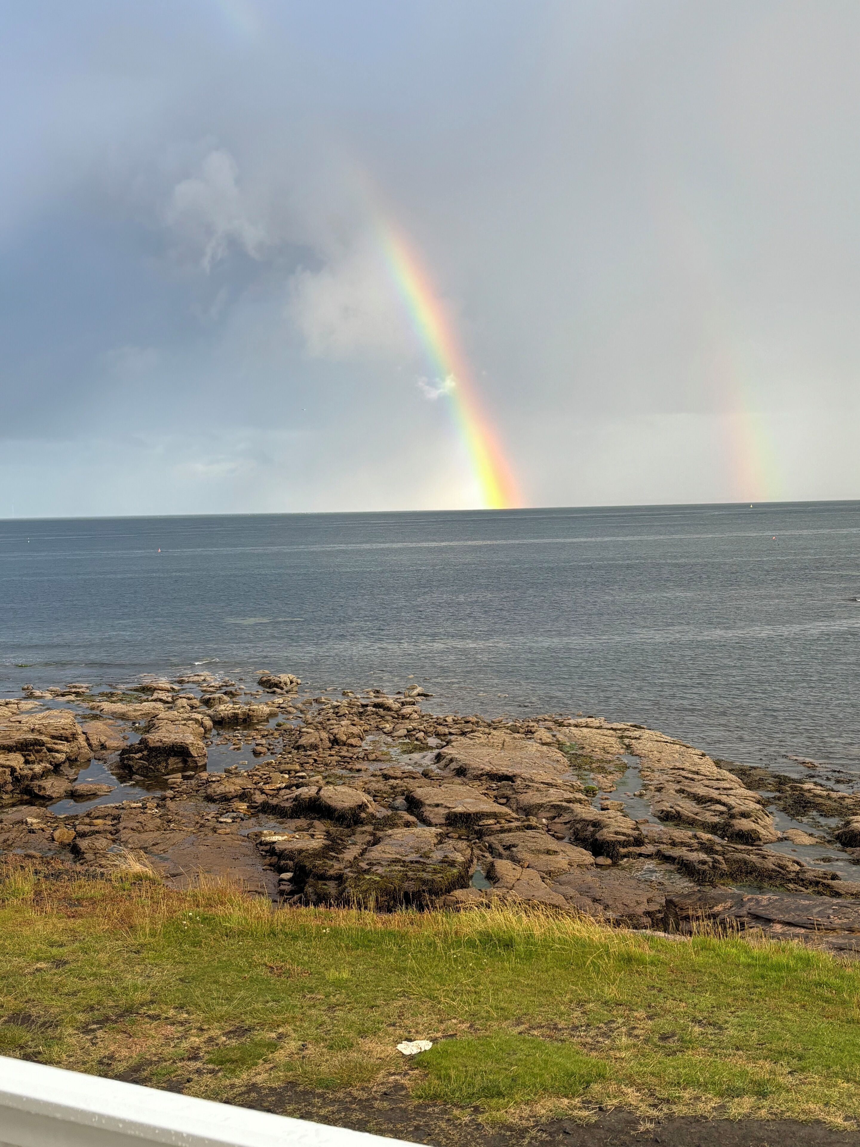 Eine Küstenlandschaft mit einem Regenbogen über dem Meer, felsigem Ufer und grünen Wiesen.