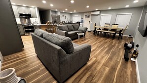 Ein modernes Wohnzimmer mit grauem Sofa, einem Essbereich mit einer Bank und einer Küche mit Edelstahlgeräten.