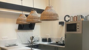 Une cuisine moderne avec un plan de travail en marbre, un four à micro-ondes encastré et une étagère en bois au-dessus de la cuisinière. On y trouve trois suspensions en osier et une petite plante en pot sur l’îlot central.