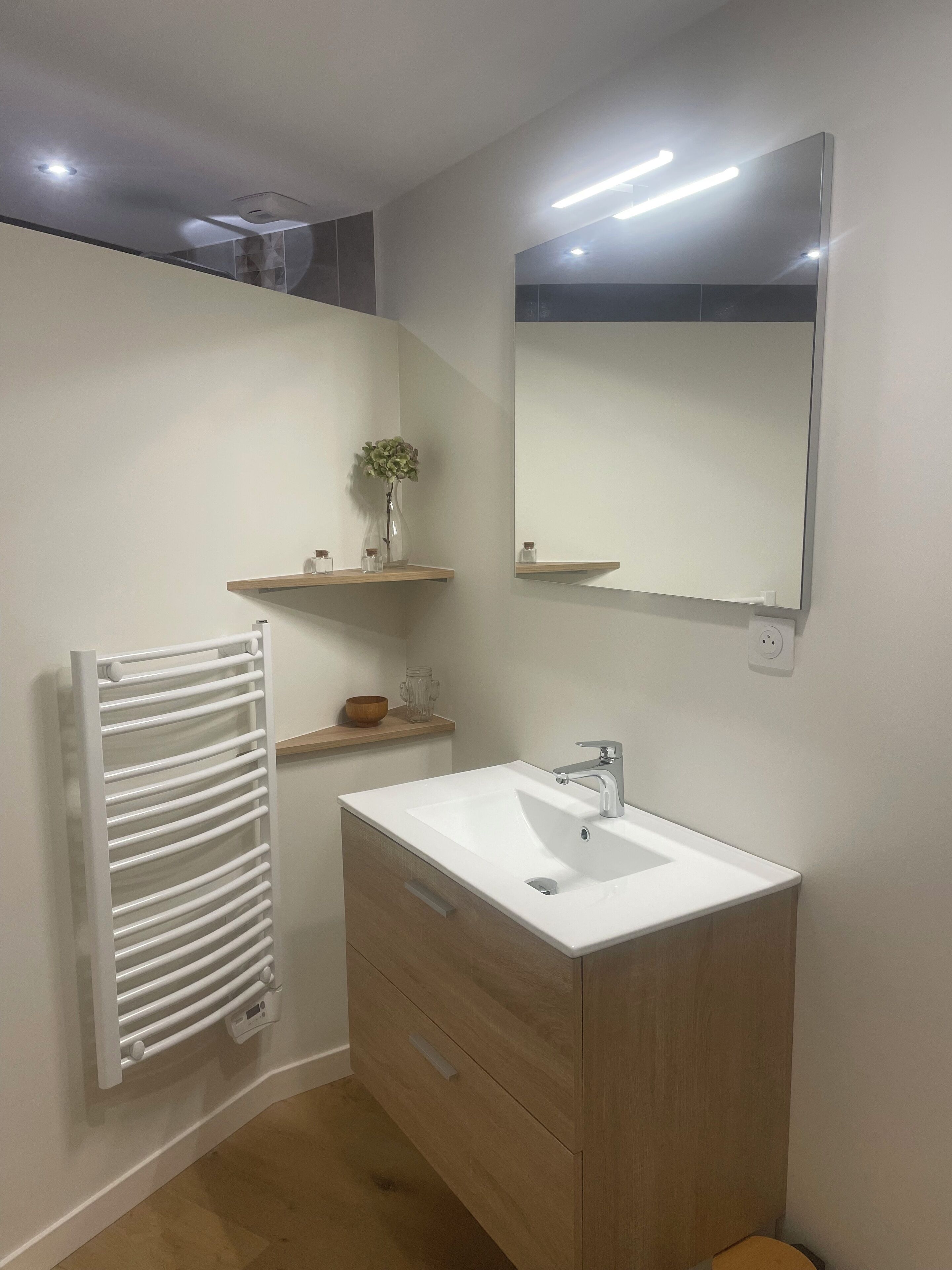 Une salle de bain moderne avec un lavabo blanc, un meuble-lavabo en bois, un porte-serviettes, un miroir et une petite étagère sur laquelle est posé un vase.