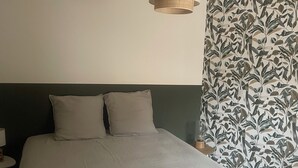Une chambre à coucher avec un lit, des oreillers, une table de chevet, une lampe et un mur recouvert d’un papier peint à motifs de feuilles.